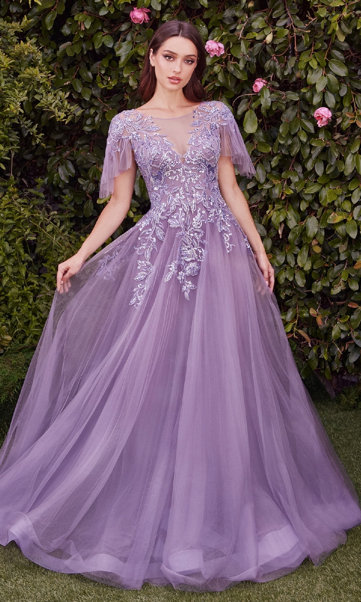 A woman stands before green leafy bushes wearing the Andrea & Leo A1351, a short-sleeve prom ball gown featuring flowing lavender fabric and sequin embroidery.